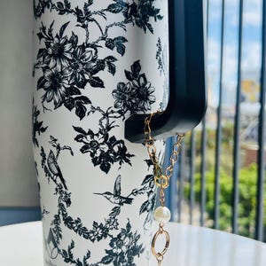 May include: White travel mug with a black floral and bird pattern. The mug has a black handle and a gold chain with two faux pearls attached. The word "wellness" is printed on the mug.