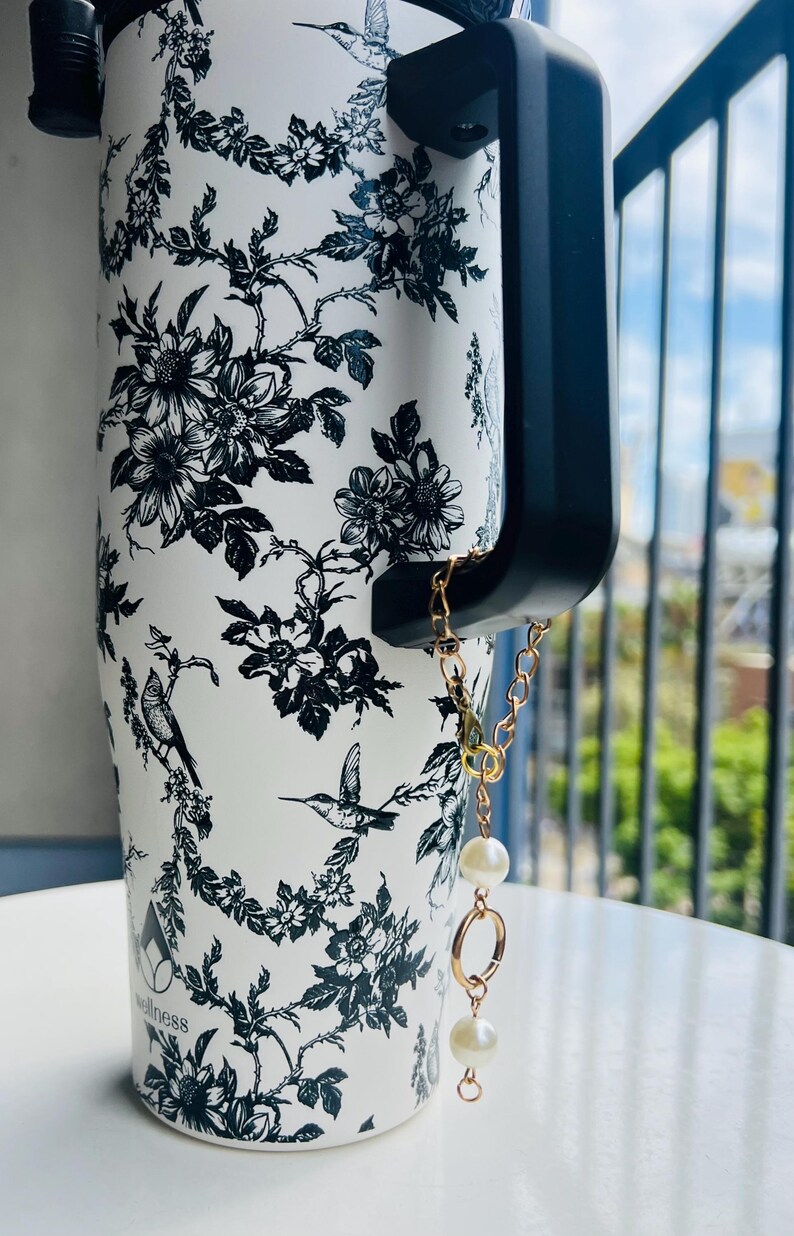 May include: White travel mug with a black floral and bird pattern. The mug has a black handle and a gold chain with two faux pearls attached. The word "wellness" is printed on the mug.