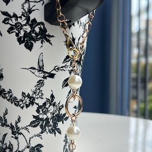 May include: A gold-colored chain with two white pearl beads and a gold ring is attached to a black object. The object is placed on a white surface. The background is a white and black floral design.