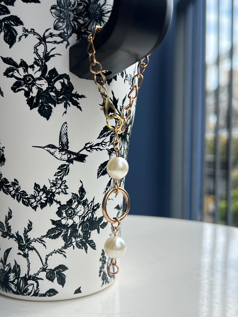 May include: A gold-colored chain with two white pearl beads and a gold ring is attached to a black object. The object is placed on a white surface. The background is a white and black floral design.