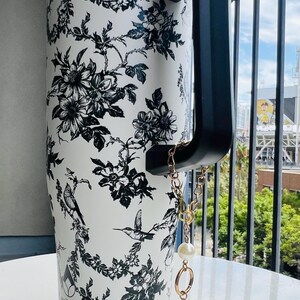May include: White tumbler with a black floral and bird pattern. The tumbler has a black handle and lid, and a gold chain with pearl accents. The word "wellness" is printed on the tumbler.