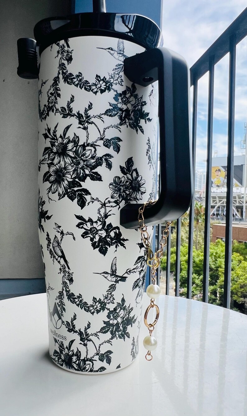 May include: White tumbler with a black floral and bird pattern. The tumbler has a black handle and lid, and a gold chain with pearl accents. The word "wellness" is printed on the tumbler.