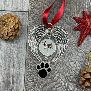 May include: Silver Christmas ornament with a paw print charm and a photo of a dog with angel wings. The ornament is hanging from a red ribbon.