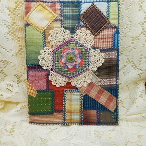 May include: A handmade patchwork journal cover with a variety of fabric squares in shades of green, red, blue, and brown. A cream-colored doily with a floral center is sewn onto the front, adding a delicate touch to the rustic design.