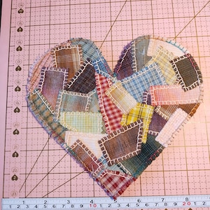 May include: A heart-shaped fabric patch made of various multicolored plaid and checkered textile scraps joined with white whipstitching. A measuring tape placed along the bottom edge shows the heart is approximately 8.5 inches wide.