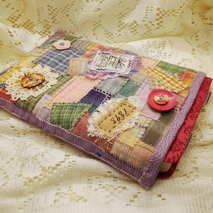 May include: A rectangular fabric book cover featuring a patchwork design of various plaid and solid colored textile scraps in shades of green, pink, yellow, and blue. The cover is embellished with lace, a small flower, a button, and a patch with the number 7898.