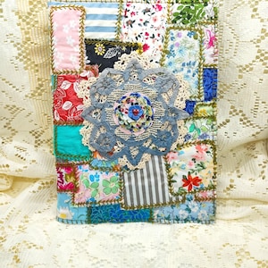 May include: A handmade journal with a patchwork cover. The cover is made of various fabric scraps in floral, striped, and solid patterns, stitched together with a decorative stitch. A large, gray lace doily is centered on the cover, with a smaller floral embellishment.