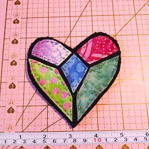 May include: A heart-shaped fabric patch featuring a stained-glass style design with multicolored patterned segments in pink, green, blue, and purple, outlined in black stitching. A measuring tape shows the patch is approximately 3.5 inches wide.