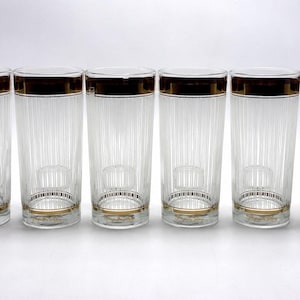 May include: A set of eight clear glass tumblers arranged in a row. Each cylindrical glass features vertical ribbed detailing along the body, a thick gold band around the top rim, and a thinner gold band accenting the base of the glass.