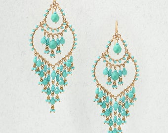 May include: A pair of gold-tone chandelier earrings featuring a teardrop-shaped frame adorned with numerous small, round, turquoise-colored beads. The design includes multiple dangling strands of beads suspended from the center and bottom edges.