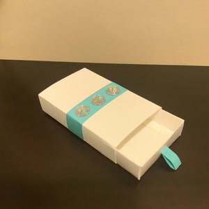 May include: A white cardboard box with a turquoise ribbon and iridescent embellishments. The box has a pull-out drawer.