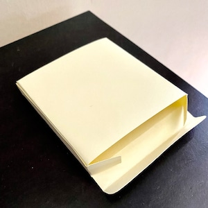 Könnte beinhalten: Eine teilweise geöffnete, rechteckige, cremefarbene Schachtel. Die Schachtel besteht aus Papier und liegt auf einer schwarzen Oberfläche. Das Innere der Schachtel ist sichtbar und zeigt einen helleren Cremeton. Das Design deutet auf eine Verpackung für kleine Gegenstände hin.