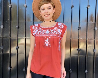 May include: A woman wearing a rust-colored embroidered blouse with white embroidery, black leggings, and a brown wide-brimmed hat. The blouse has a scoop neck and short sleeves. The woman is standing in front of a black metal gate.