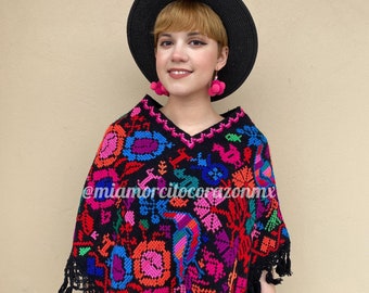 May include: A black poncho with colorful embroidery featuring floral and bird designs. The poncho has fringe around the edges and is worn by a person with short blonde hair wearing a black hat and pink pom-pom earrings. The text "@miamorcilocoazonmx" is visible on the poncho.