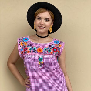 Peut inclure: Un chemisier sans manches en vichy rose et blanc avec des broderies florales colorées. Le chemisier a un col rond et une coupe ample. Le mannequin porte un chapeau noir et un short noir.