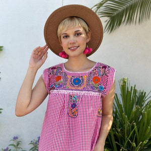 Puede incluir: Una blusa de cuadros vichy rosa y blanco con bordados coloridos alrededor del escote. La blusa tiene mangas cortas y un escote redondo. La modelo lleva un sombrero marrón y pendientes de borla rosa.