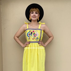 May include: A yellow midi dress with a colorful embroidered design on the bodice. The dress has thin straps that tie at the shoulders and a black trim around the neckline. The dress has a black and pink trim at the hem.