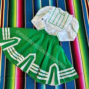Puede incluir: Una falda verde con ribete de cinta blanca y una blusa blanca con bordados verdes. La falda y la blusa están sobre una manta de rayas de colores.