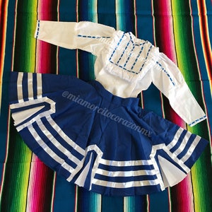 Puede incluir: Una falda azul con ribete de cinta blanca y una blusa blanca con bordados azules. La falda y la blusa están sobre una manta de rayas de colores.