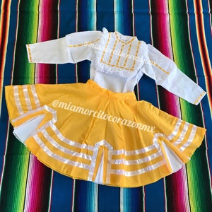 May include: A yellow skirt with white ribbon trim and a white blouse with yellow embroidery. The skirt has a ruffled hem and the blouse has a ruffled collar. The outfit is on a colorful striped blanket.