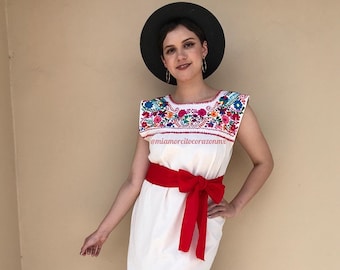 May include: A white maxi dress with colorful embroidery around the neckline and a red sash tied at the waist. The dress is worn by a person wearing a black hat and has the text "@miamorcilocorazonn" on the front.