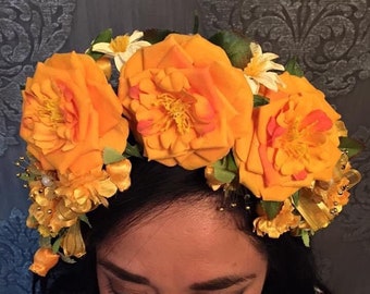 May include: A woman wearing a white embroidered blouse with a floral pattern and a yellow flower crown. The crown is made of fabric flowers and has a few small pearl accents.