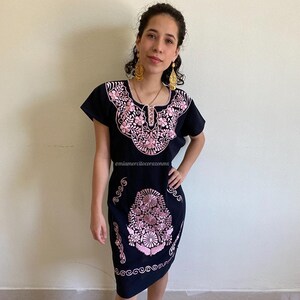 May include: A black embroidered dress with pink floral designs. The dress has short sleeves and a scoop neckline. The model is wearing the dress with brown sandals and gold earrings. The text "@miamorcilcorazonmx" is visible on the dress.