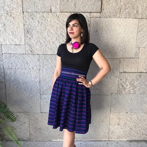 May include: A woman wearing a black short-sleeved shirt with a pink and blue necklace. She is wearing a black and purple striped skirt with a black waistband that says "@miamordilecerrania.mx". She is wearing black heeled sandals.