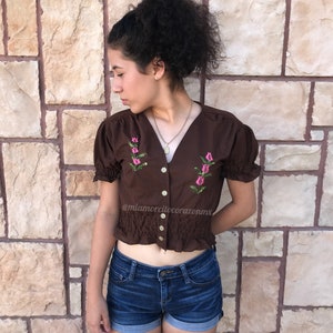 May include: A brown cropped top with short sleeves and a button-up front. The top has pink embroidered flowers and a ruffled hem. The model is wearing blue denim shorts.