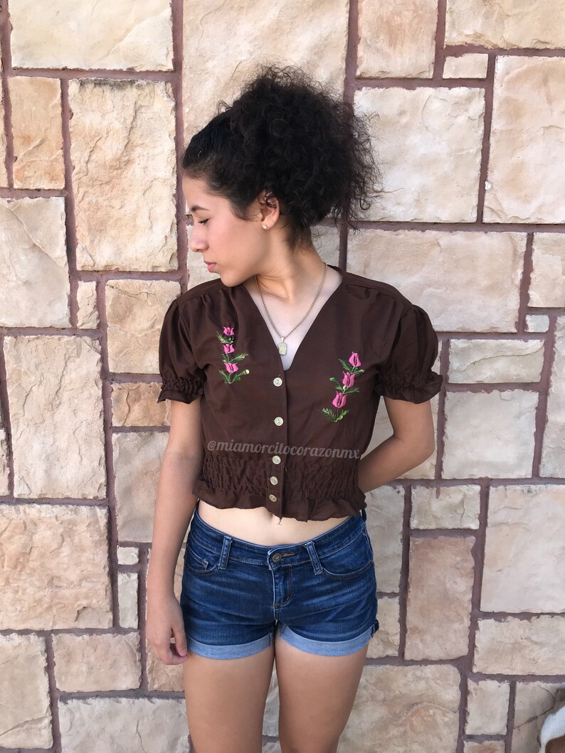 May include: A brown cropped top with short sleeves and a button-up front. The top has pink embroidered flowers and a ruffled hem. The model is wearing blue denim shorts.