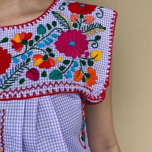 Peut inclure: Un haut sans manches en vichy violet et blanc avec des fleurs brod&eacute;es color&eacute;es. La broderie pr&eacute;sente une vari&eacute;t&eacute; de fleurs dans des tons de rouge, rose, orange, jaune, bleu et vert.