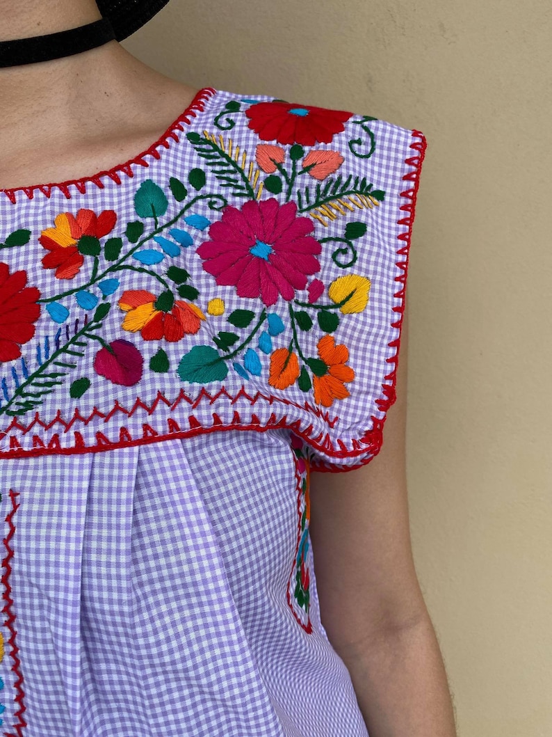 Peut inclure: Un haut sans manches en vichy violet et blanc avec des fleurs brod&eacute;es color&eacute;es. La broderie pr&eacute;sente une vari&eacute;t&eacute; de fleurs dans des tons de rouge, rose, orange, jaune, bleu et vert.