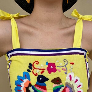 May include: Yellow sleeveless top with a colorful embroidered design of birds and flowers. The top has yellow straps that tie into bows at the shoulders.