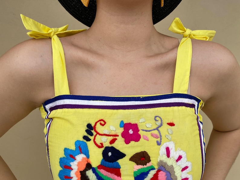 May include: Yellow sleeveless top with a colorful embroidered design of birds and flowers. The top has yellow straps that tie into bows at the shoulders.