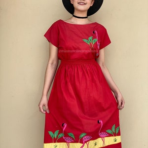 May include: A red midi dress with short sleeves and a gathered waist. The dress features embroidered pink flamingos and green palm leaves on the skirt. The dress has a yellow band at the hemline. The model is wearing a black hat and a black choker.
