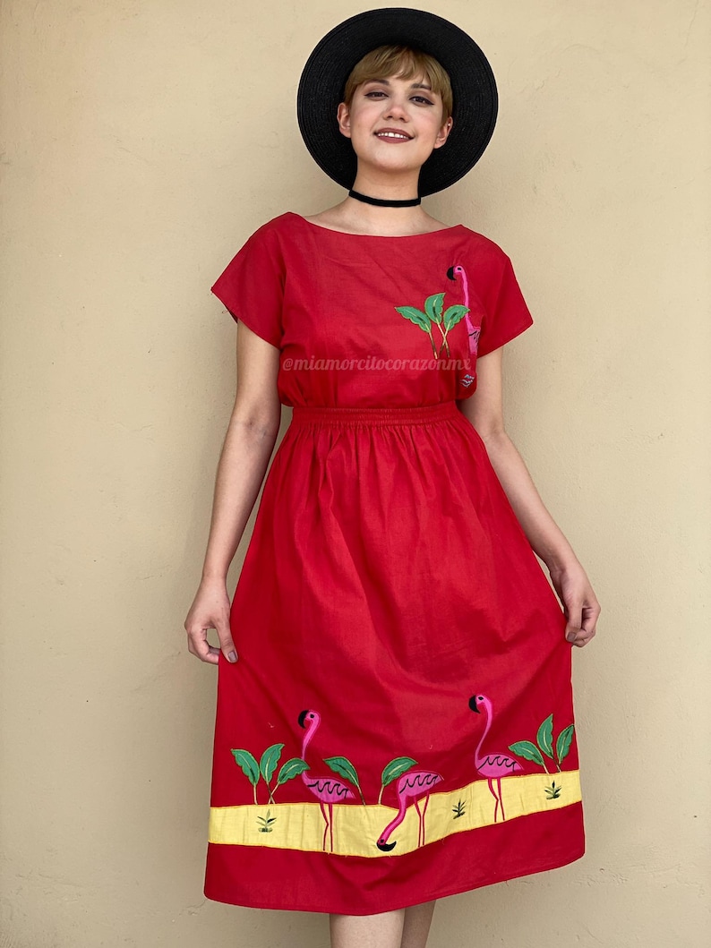 May include: A red midi dress with short sleeves and a gathered waist. The dress features embroidered pink flamingos and green palm leaves on the skirt. The dress has a yellow band at the hemline. The model is wearing a black hat and a black choker.