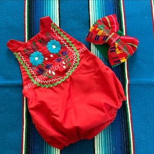 May include: Red baby romper with floral embroidery in blue, purple, and green. The romper has a matching bow with colorful stripes and patterns. The outfit is displayed on a striped textile background.