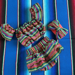 May include: A colorful, striped baby outfit set, including a top, shorts, and a bow. The fabric features horizontal stripes in shades of red, green, yellow, and blue. The set is displayed on a blue and striped textile background.