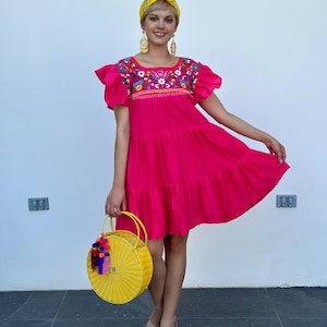 May include: A bright pink, tiered dress with embroidered floral details on the bodice and short, ruffled sleeves. A yellow woven handbag with tassel accents and a yellow headband complete the look. The text "@inmiamordecorazon" is embroidered on the dress.