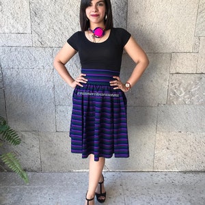 May include: A woman wearing a black top and a skirt with purple, green, and white stripes. She is wearing a pink and black necklace and has her hair pulled back. The skirt is a midi length and has a high waist. The woman is standing in front of a stone wall.