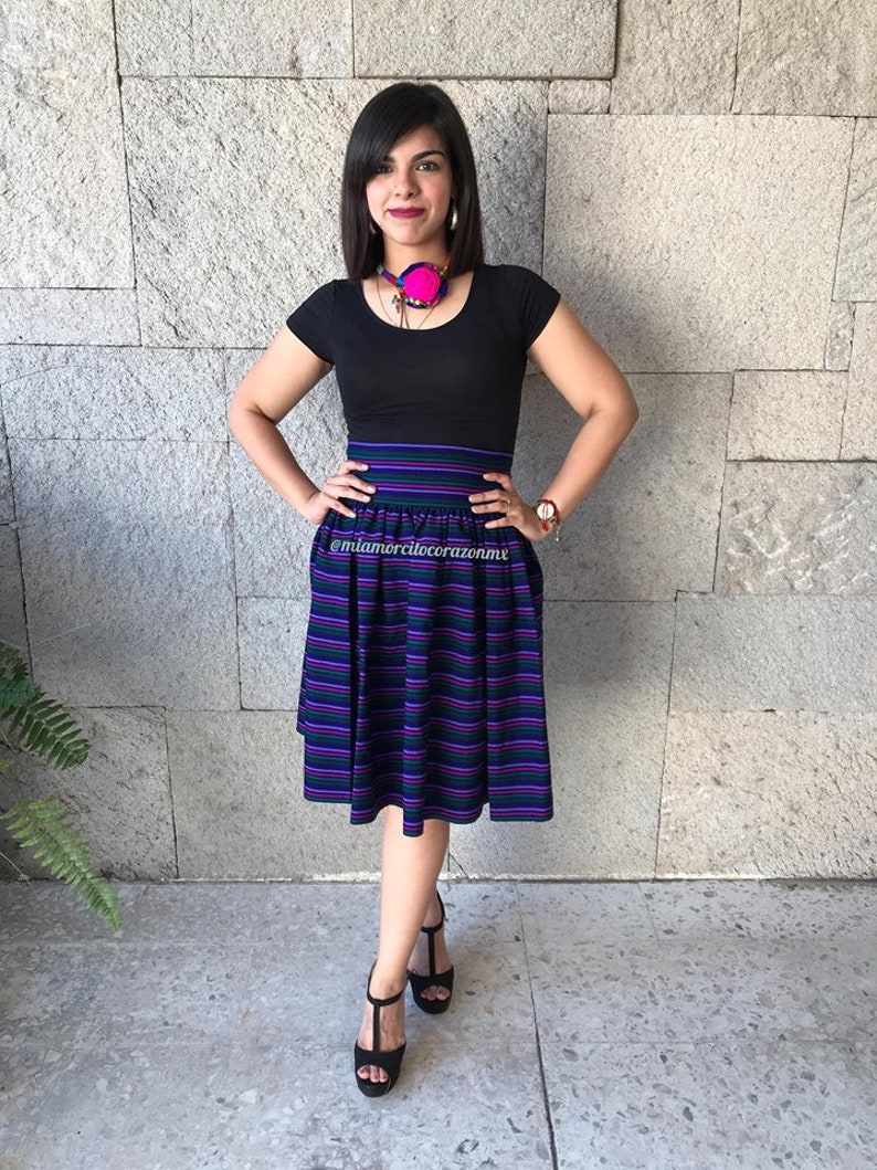 May include: A woman wearing a black top and a skirt with purple, green, and white stripes. She is wearing a pink and black necklace and has her hair pulled back. The skirt is a midi length and has a high waist. The woman is standing in front of a stone wall.
