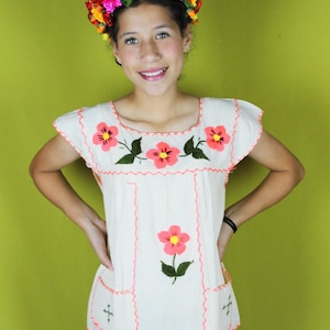 May include: A young person wearing a white embroidered blouse with pink flowers and green leaves. The blouse has short sleeves and a square neckline. The person is also wearing a flower crown with red, yellow, and orange flowers. The background is a solid green.