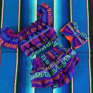Könnte beinhalten: Ein lila und blaues Outfit mit einem farbenfrohen geometrischen Muster. Das Outfit besteht aus einem Oberteil mit Puffärmeln und einem Rock mit mehreren Rüschen. Ein passendes Stirnband mit einer großen Schleife ist im Lieferumfang enthalten.