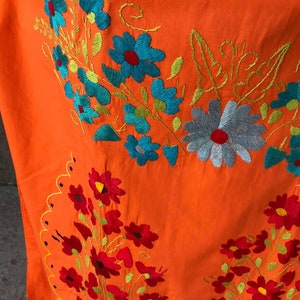 May include: Orange fabric with embroidered floral designs in shades of red, blue, and green. The flowers are arranged in clusters and have a vintage style.