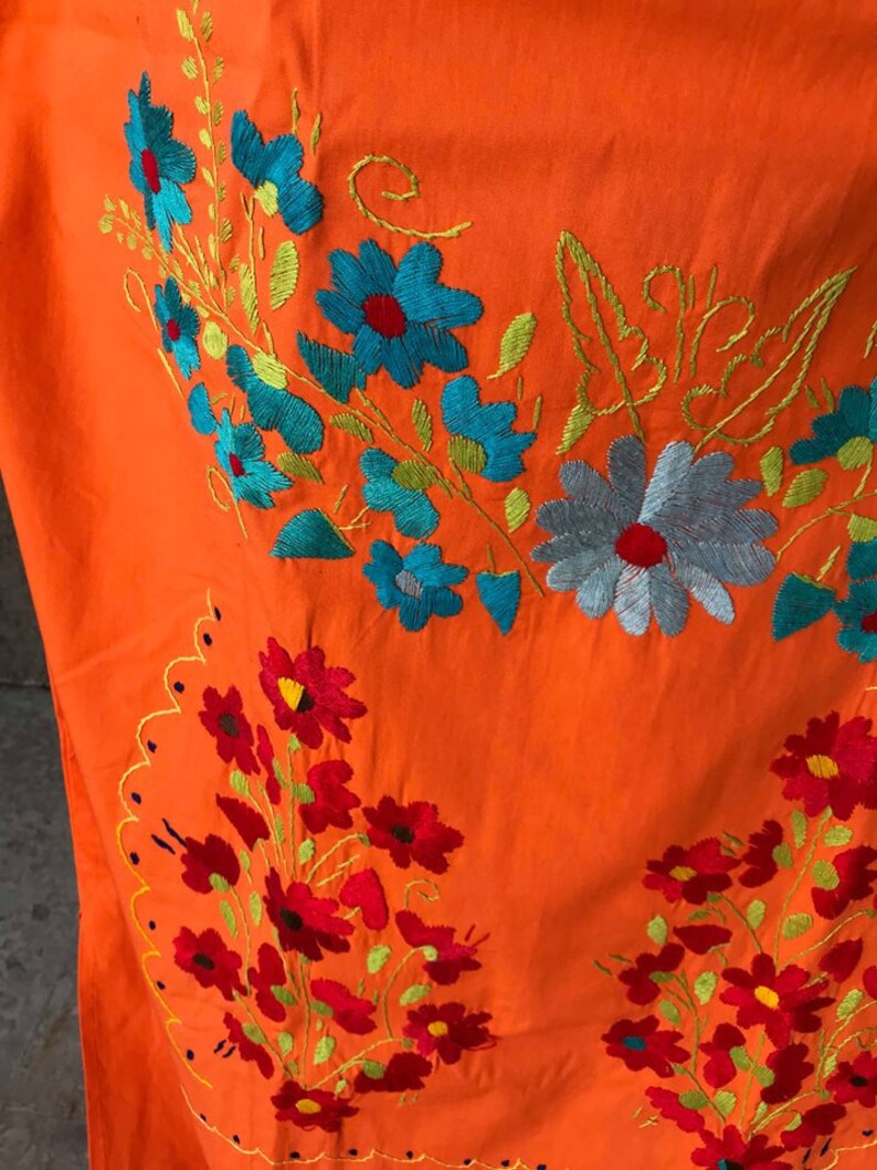 May include: Orange fabric with embroidered floral designs in shades of red, blue, and green. The flowers are arranged in clusters and have a vintage style.