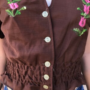 May include: A brown, short-sleeved, button-up blouse with a gathered waist and floral embroidery. The blouse has a V-neckline and features white buttons.