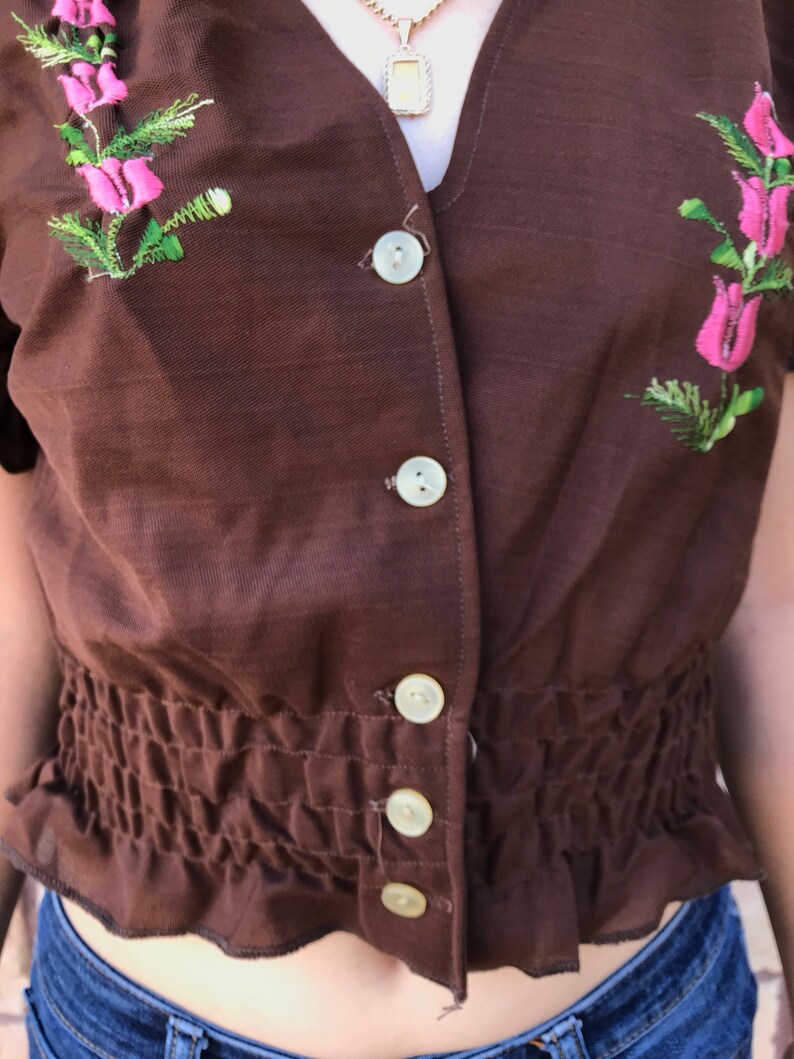 May include: A brown, short-sleeved, button-up blouse with a gathered waist and floral embroidery. The blouse has a V-neckline and features white buttons.