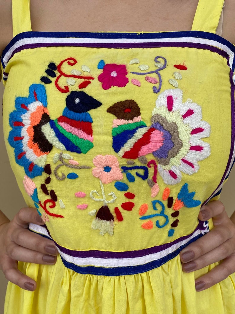 May include: Yellow sleeveless dress with colorful embroidered birds and flowers. The dress has a purple, white, and blue trim around the neckline and hem.