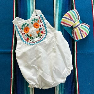 May include: White baby romper with orange floral embroidery and a matching striped bow headband. The romper features a round neckline with a floral design and a ruffled hem. The headband has a striped pattern in various colors.