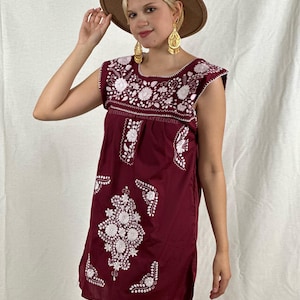 May include: A burgundy sleeveless dress with white embroidered floral designs. The dress has a round neckline and a short length. The model is wearing a brown hat and gold earrings.